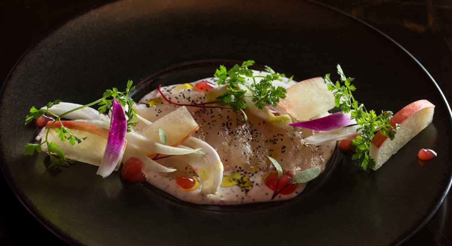 Close up view of a crudo dish decorated with sliced apples and cilantro on a black porcelain plate