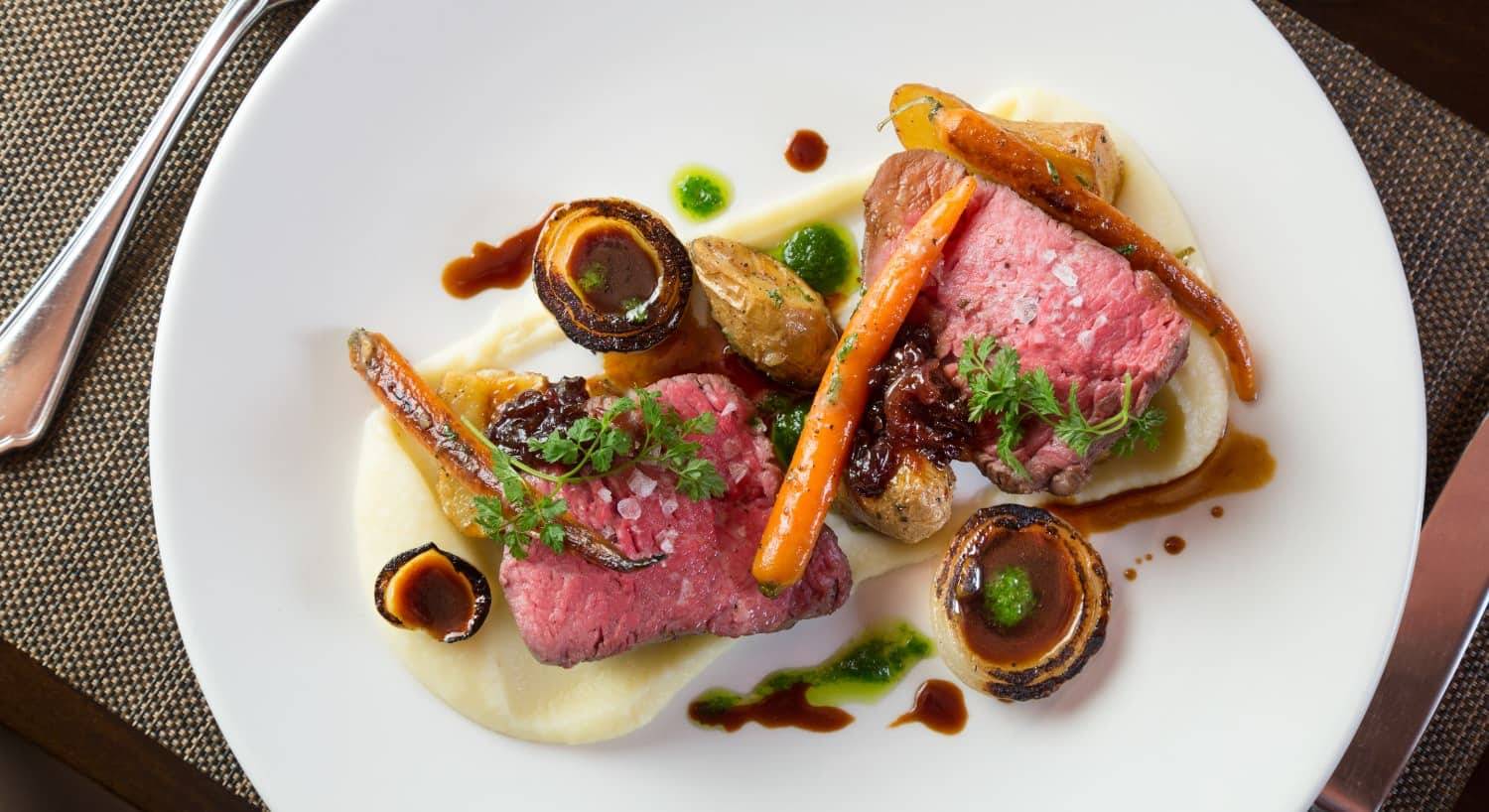 Close up view of dish with beef and roasted potatoes, carrots, and onions on white porcelain plate