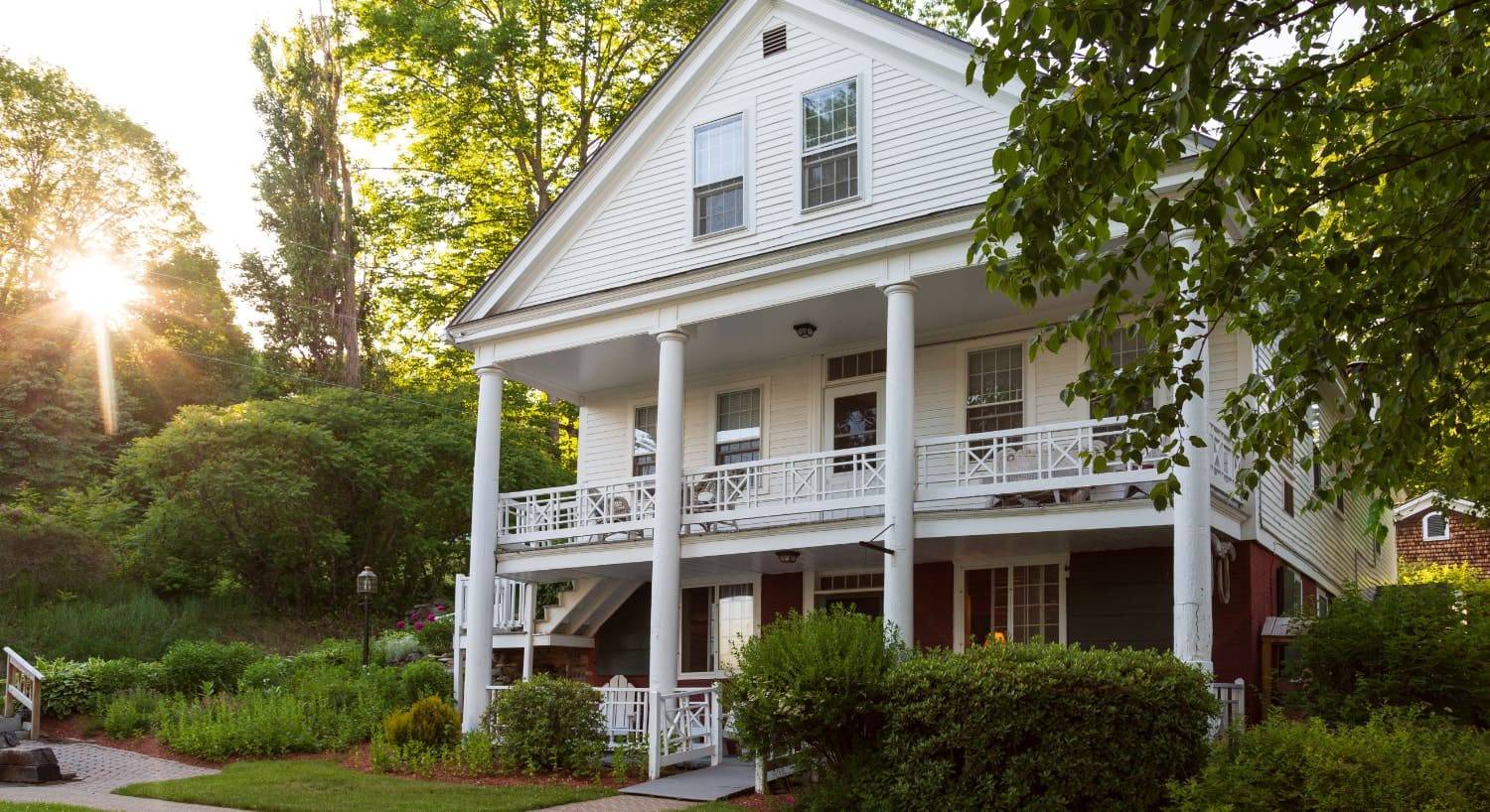 Exterior view of the property painted white with front porch and second level porch all surrounded by large green trees and shrubs with sun in the background