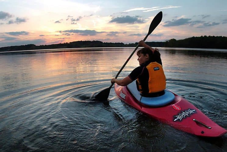 Woman wearing orange life jacket kayaking on the water with rolling hills in the background at dusk