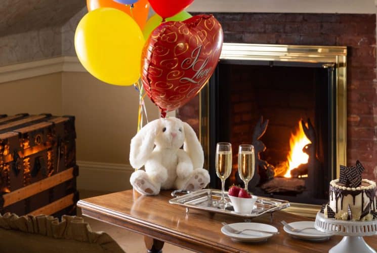 White bunny with multicolored balloons sitting on wooden coffee table with champagne-filled glasses, strawberries, and fancy dessert