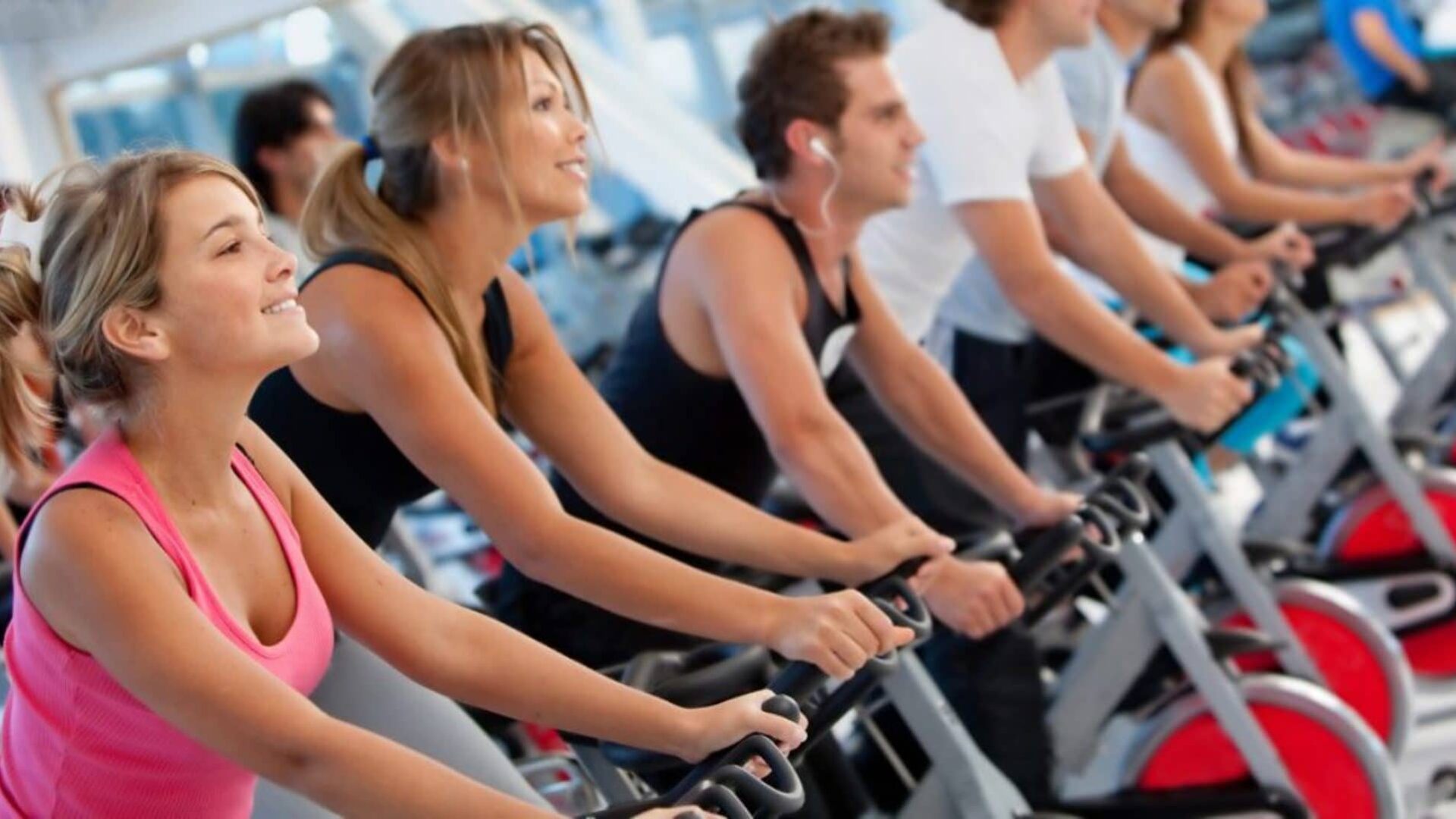 group of people riding indoor bikes in a spin class|five people on a row of treadmills at the gym