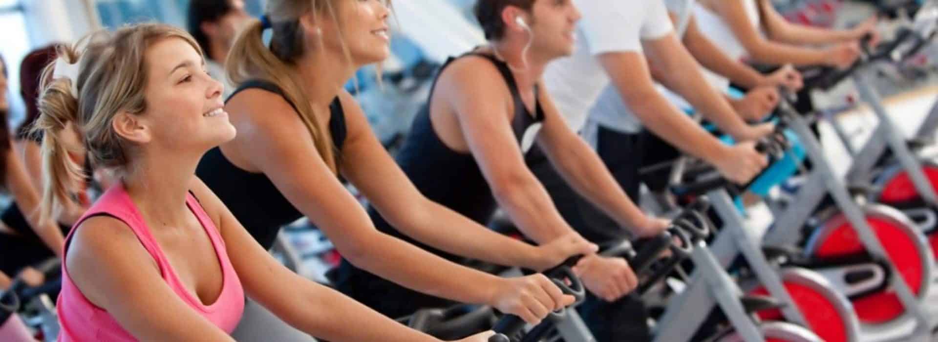 group of people riding indoor bikes in a spin class|five people on a row of treadmills at the gym